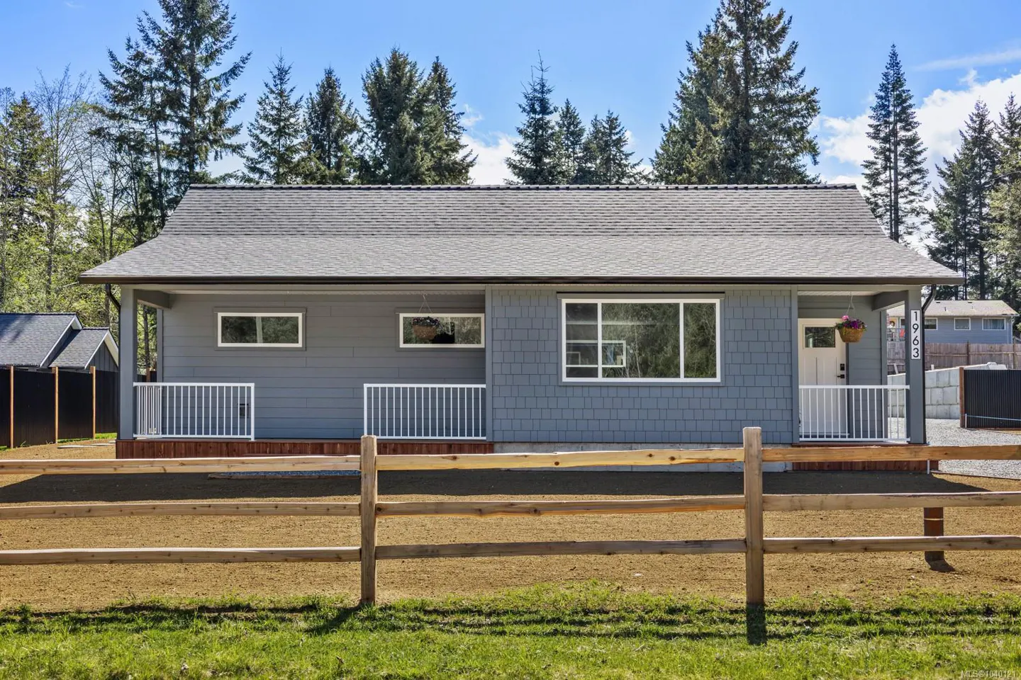 A grey, single-story house with a white porch railing and a wooden fence in the foreground, set against a backdrop of trees.