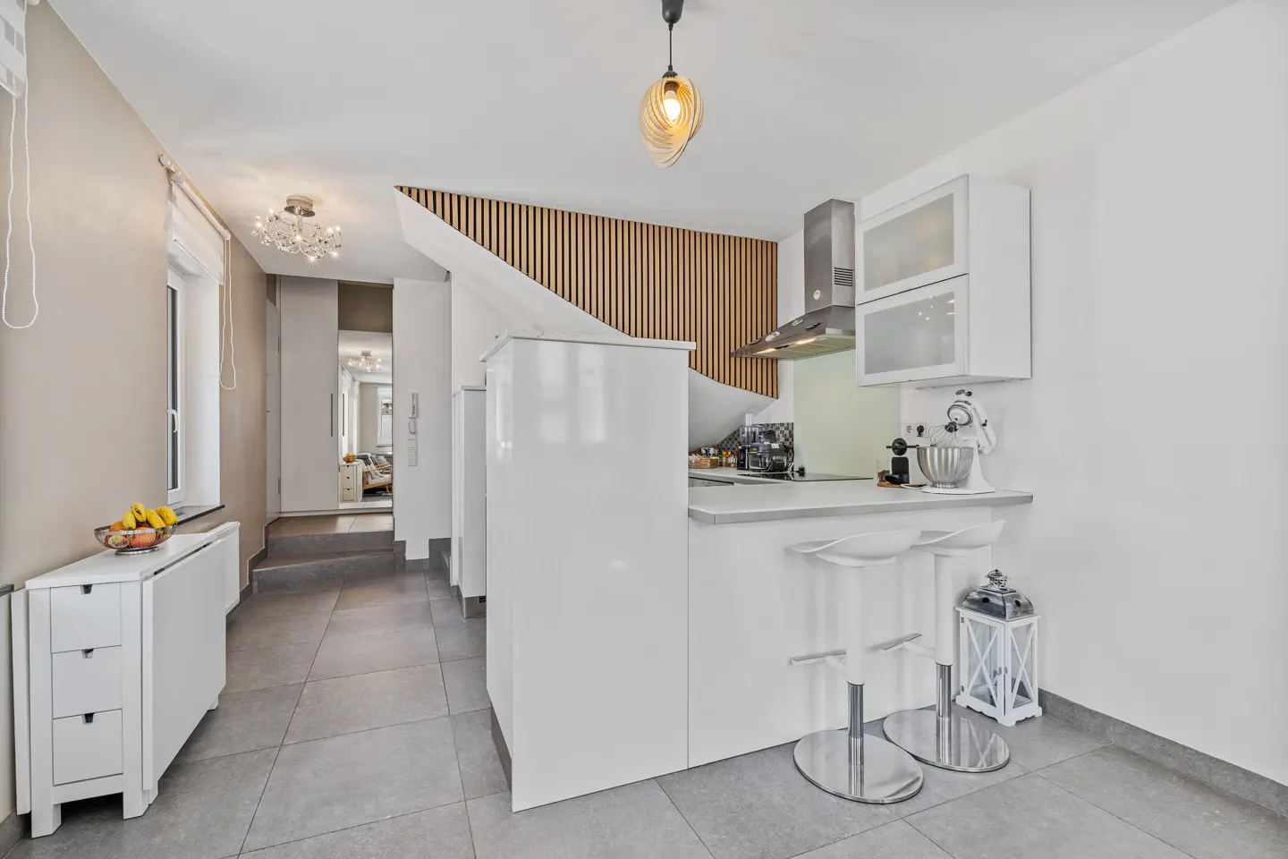 A modern kitchen with white cabinets, a breakfast bar with two clear stools, and a wooden slat wall accent. A hallway leads to another room.