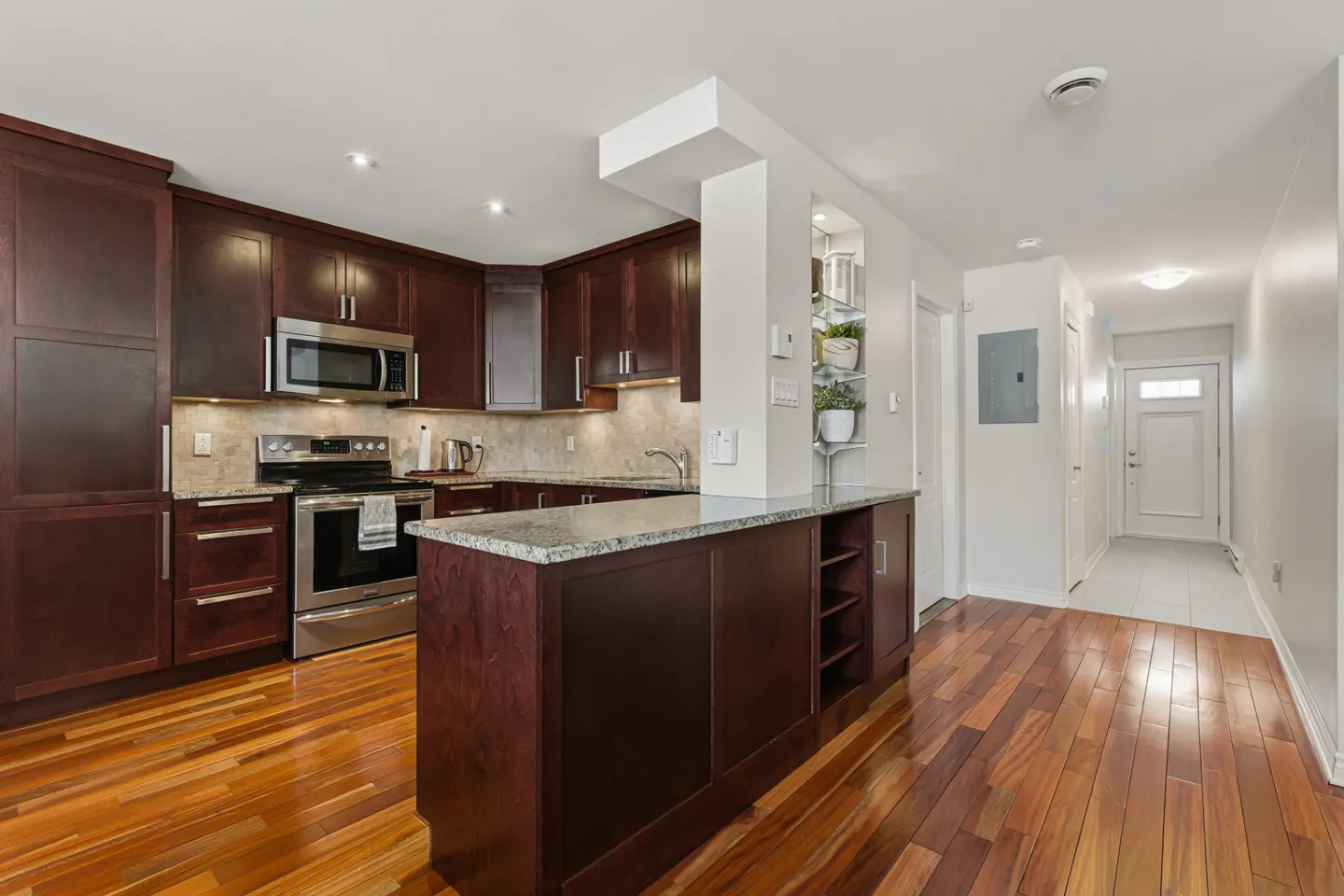 A kitchen with dark wood cabinets, stainless steel appliances, and a granite countertop island. Hardwood floors lead to a white hallway with a door.