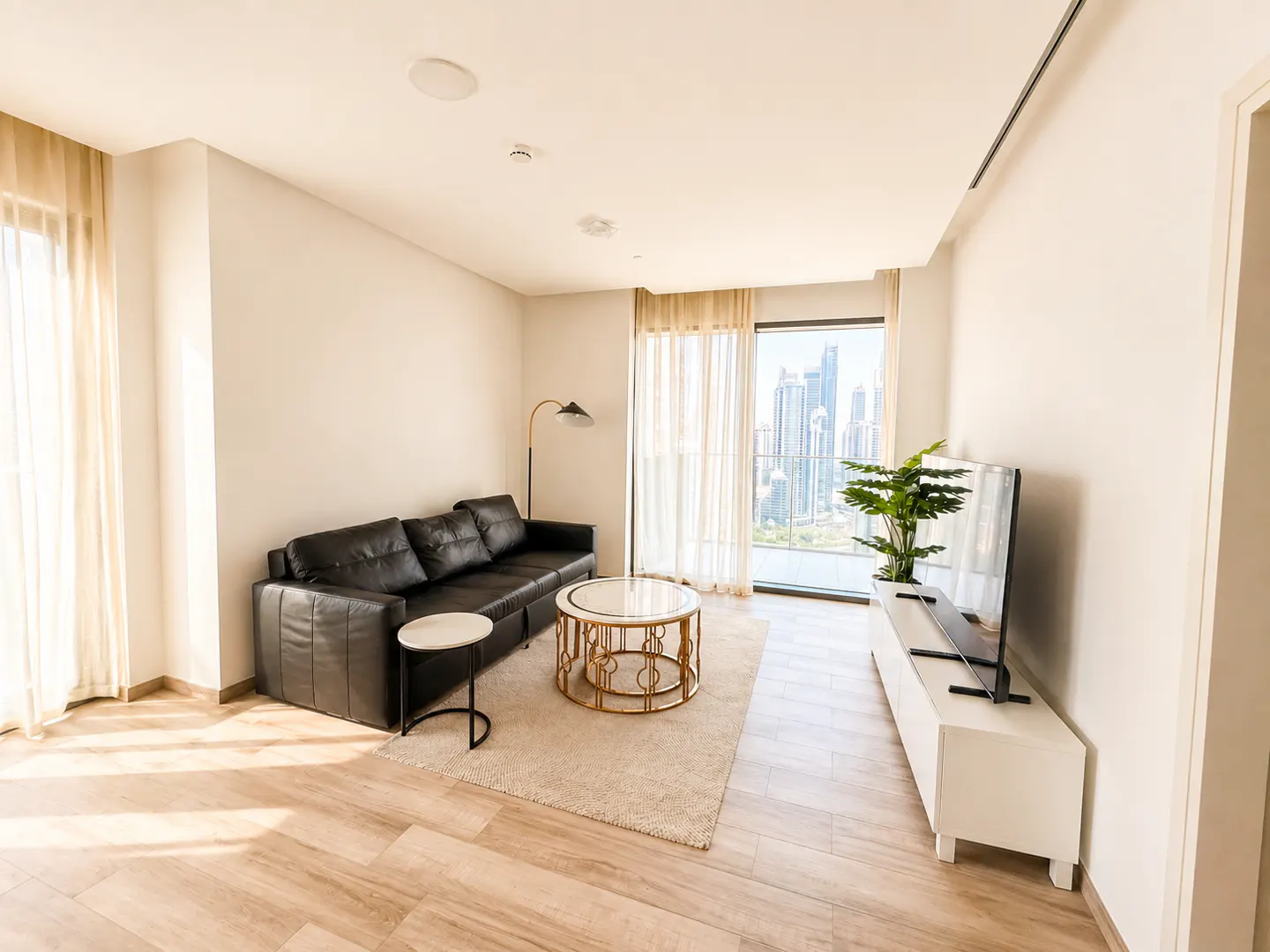 Bright living room with a black leather sofa, gold coffee table, TV, and a city view through large windows.