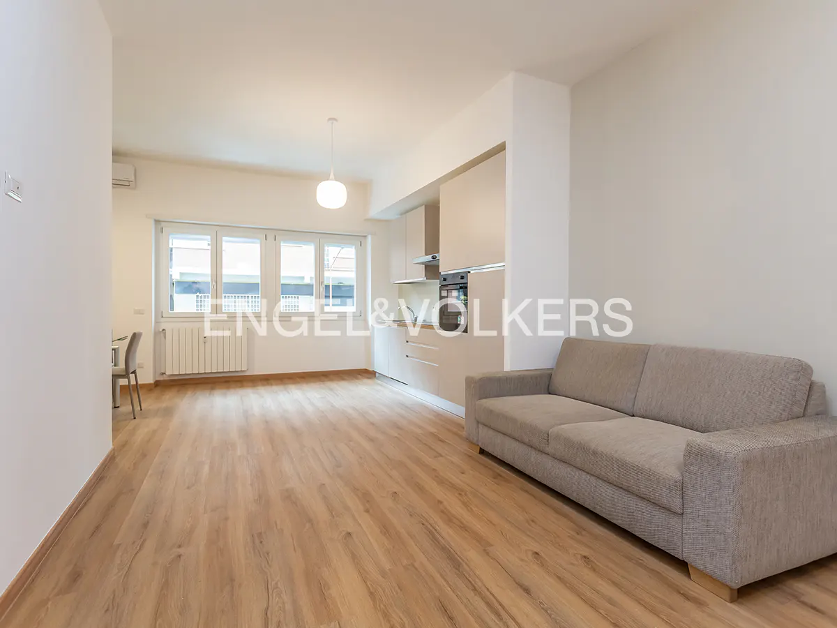 Bright, open-plan apartment with wood floors, a beige sofa, and a modern kitchen with white cabinets. Large windows provide natural light.