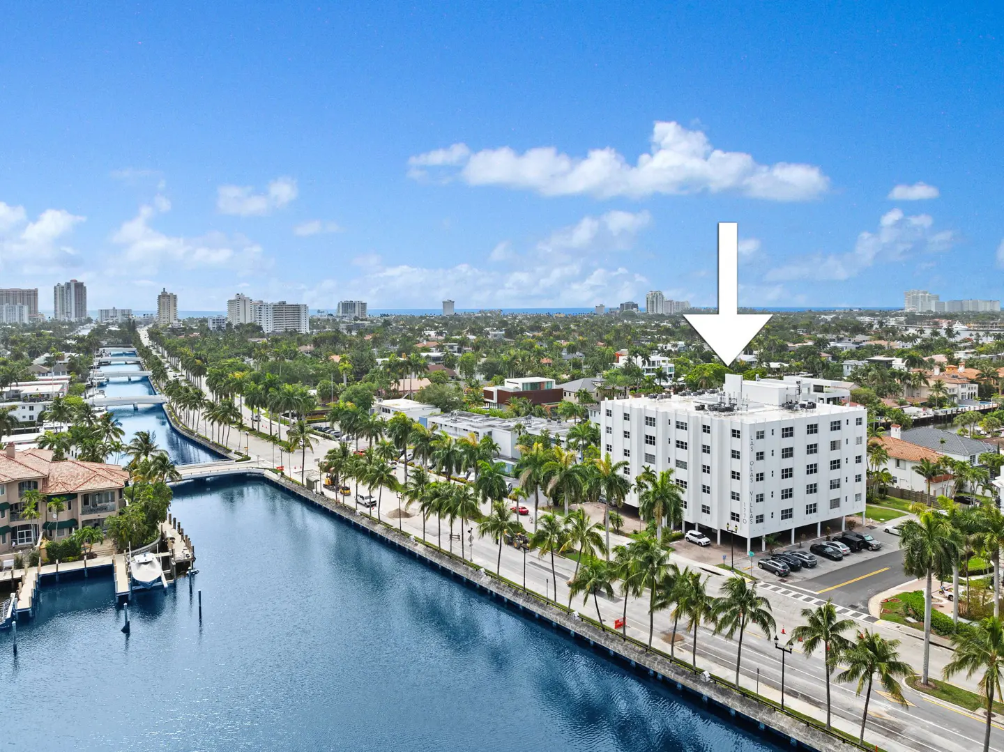 Aerial view of a white apartment building with palm trees, a canal, and a blue sky. An arrow points to the building.
