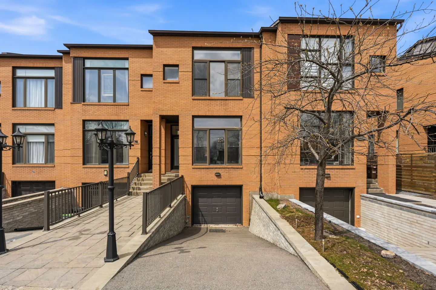 Three-story brick townhouses with dark trim, garages, and a ramp leading to the front doors. A bare tree stands in front.