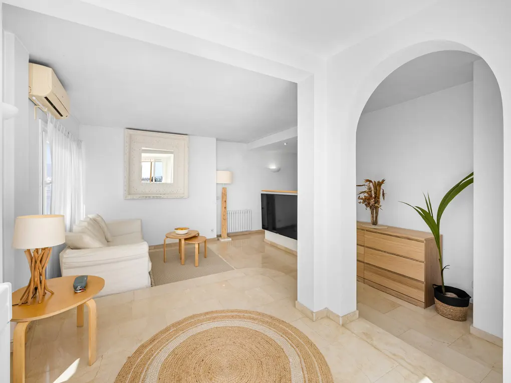 Bright, white living room with a jute rug, cream sofa, and light wood furniture. An arched doorway leads to another room.