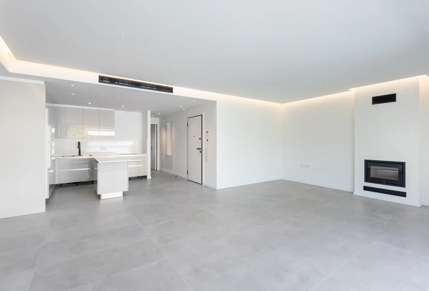 Bright, modern apartment with white walls, gray tile floor, and recessed lighting. A white kitchen and black fireplace add contrast.