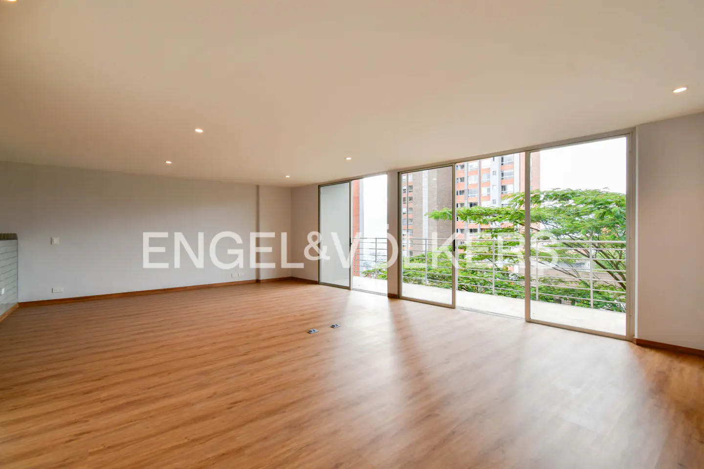 Unfurnished living room with wood floors and white walls. Large sliding glass doors open to a balcony with city views. Engel & Völkers logo on wall.