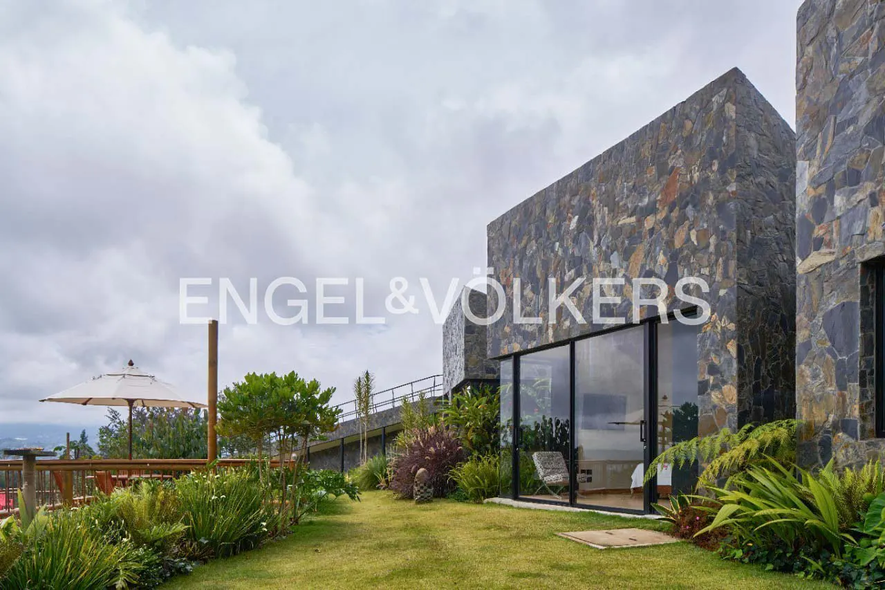 Modern stone-clad home with floor-to-ceiling glass walls, lush garden, and patio area under a cloudy sky.