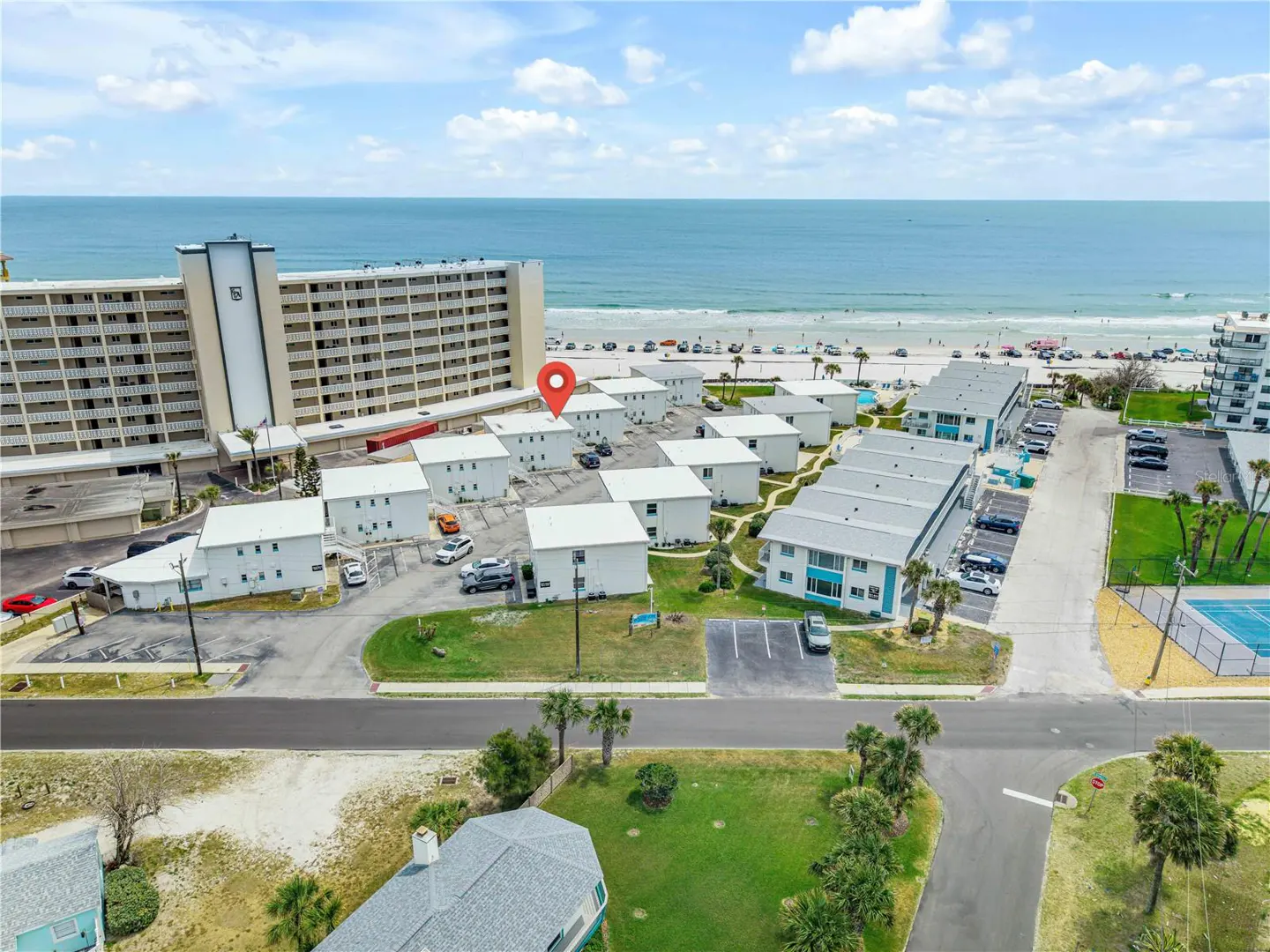 Aerial view of white beach condos with a red pin marker, near a sandy beach and blue ocean under a partly cloudy sky.