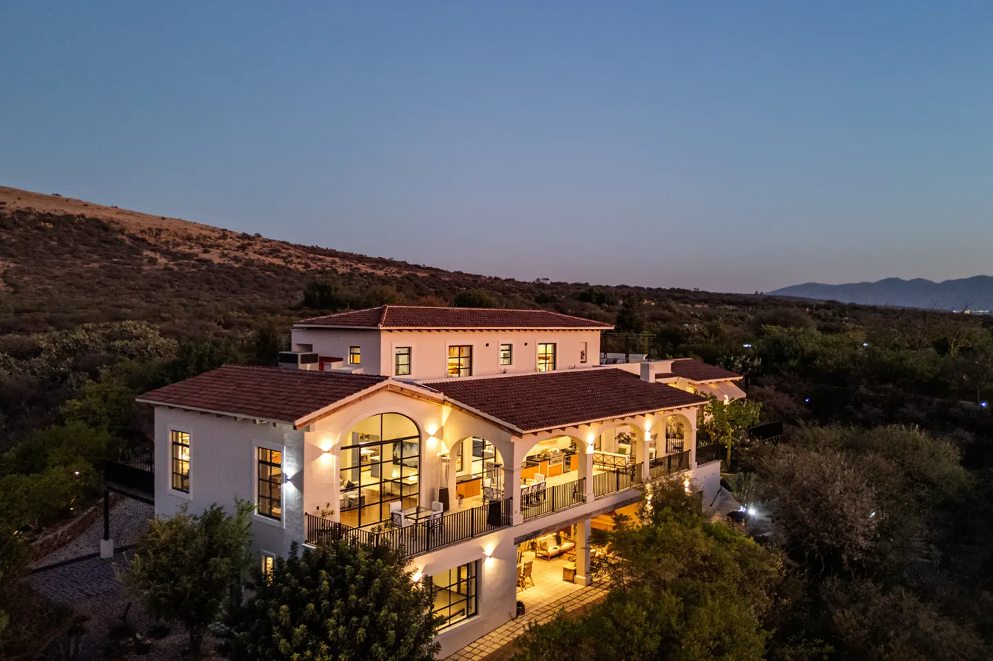 Two-story white house with a red tile roof, surrounded by trees and a hill in the background at dusk.