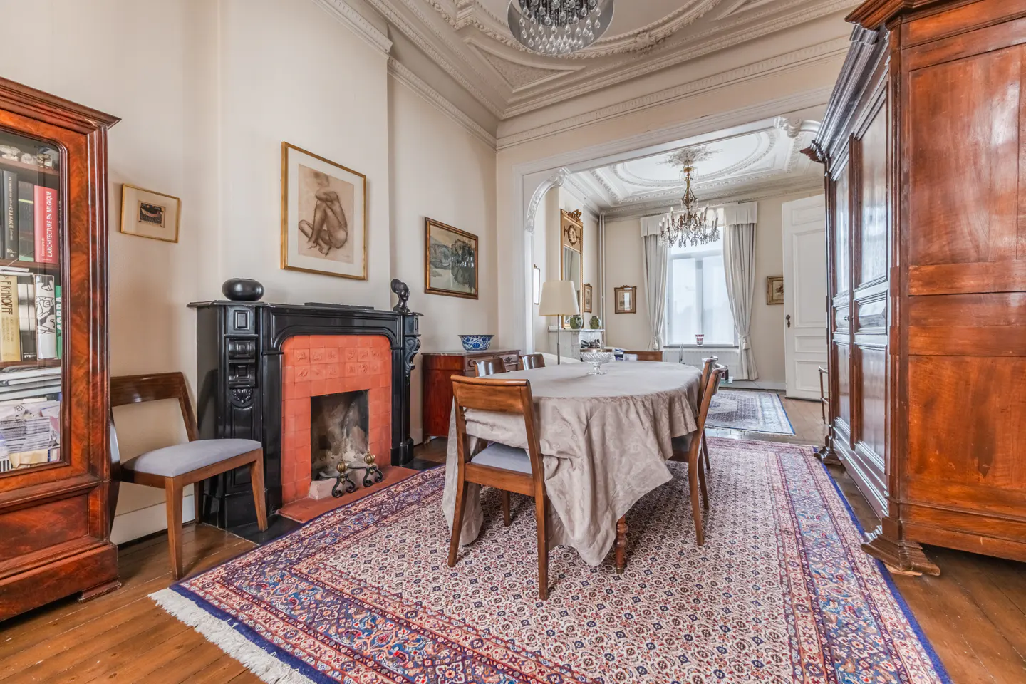 Ornate dining room with a fireplace, wood furniture, and a patterned rug. A chandelier hangs from the decorative ceiling.