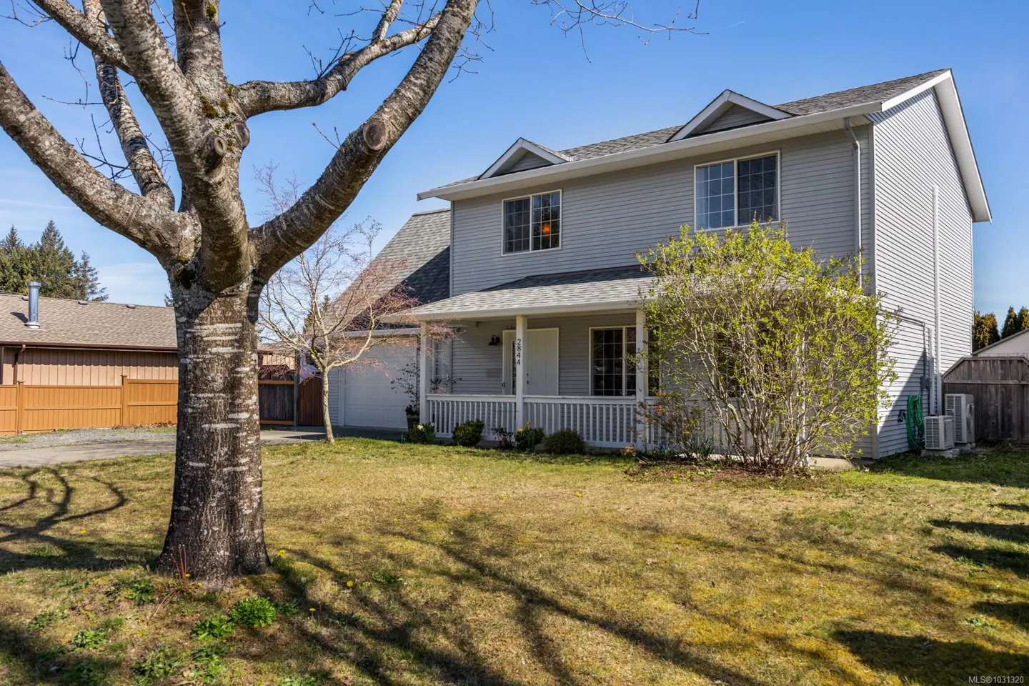 Two-story light gray house with a white porch and a green lawn. A large tree is on the left.
