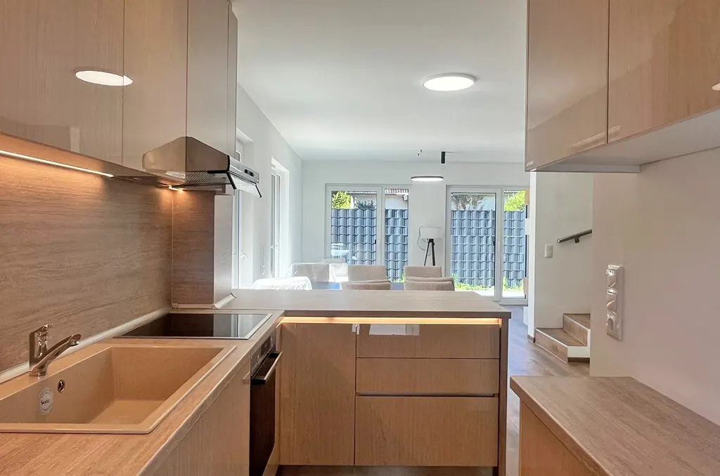 A modern kitchen with light wood cabinets, a beige sink, and a view into a bright living room.