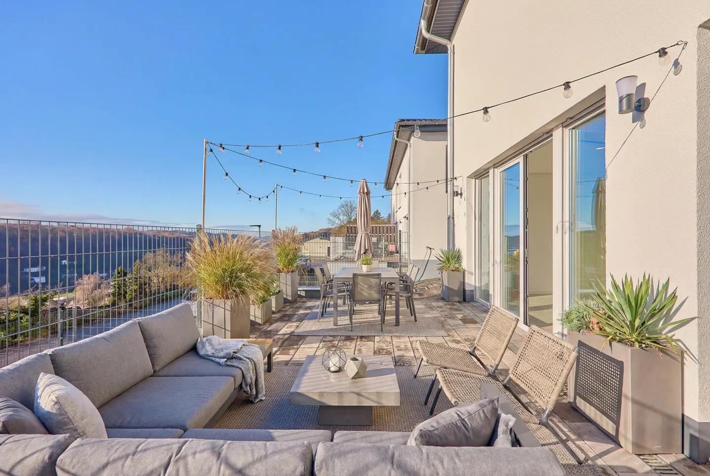 Outdoor patio with gray sectional, dining table, and string lights. A view of trees and a blue sky are in the background.