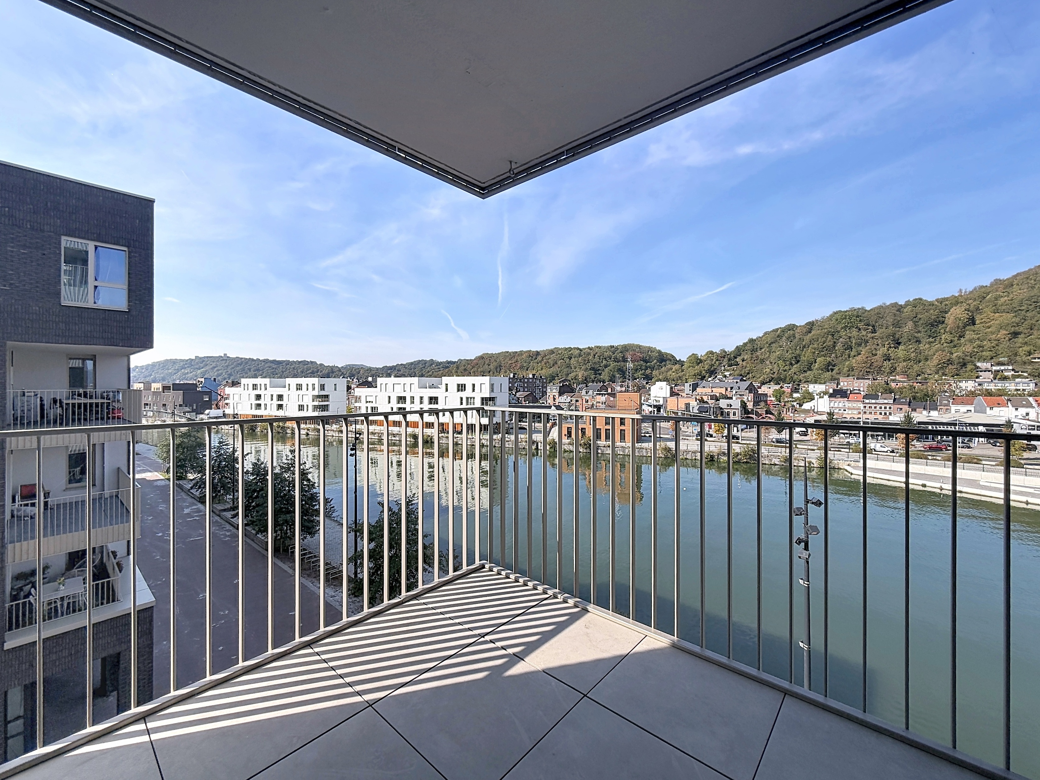 View from a modern apartment balcony overlooking a river, a tree-lined path, and a town nestled against a green hillside. [AI-generated text]
