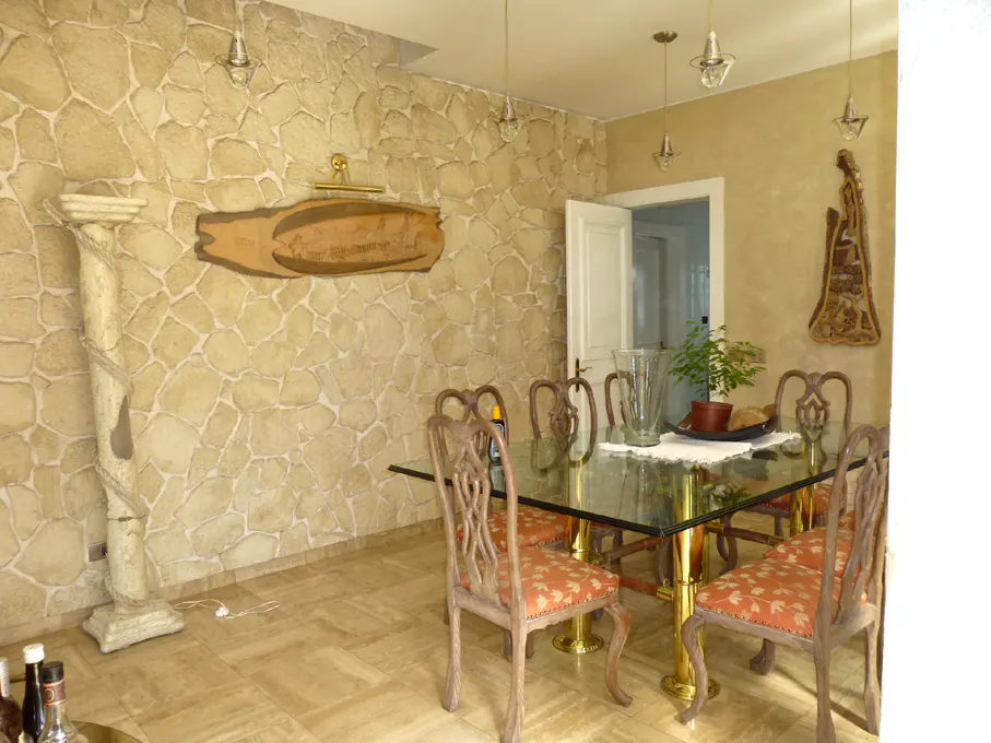 Dining room with glass table, gold legs, and floral chairs. Stone wall with column and wooden sign. Open white door in background.