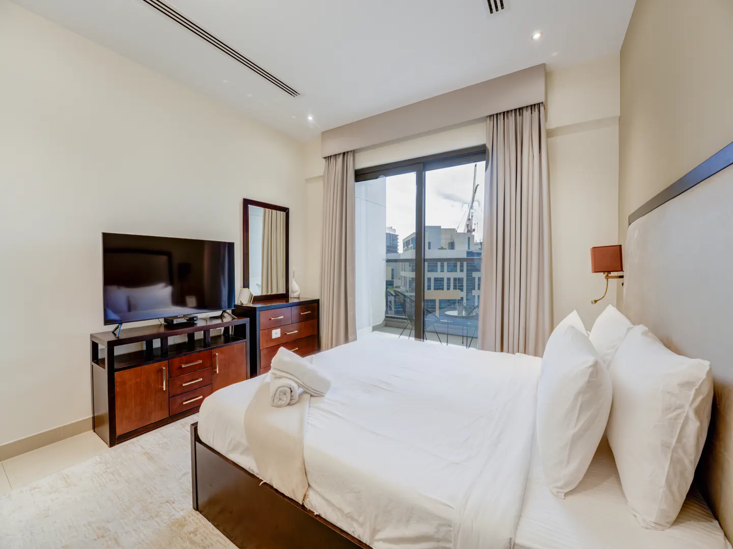 Bedroom with a white bed, TV, and balcony. Neutral walls, wood furniture, and city view. Curtains frame the sliding glass door.