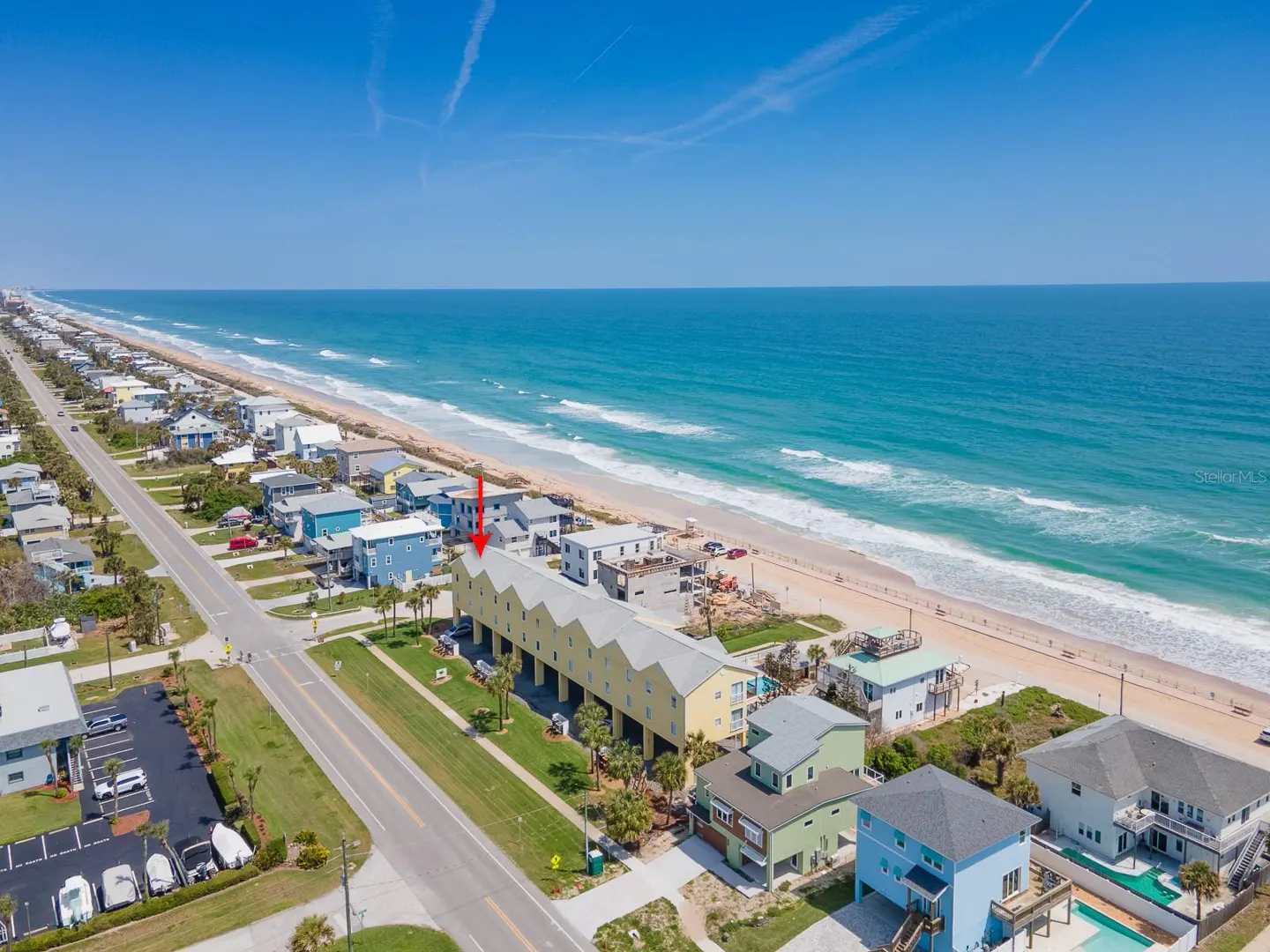 Aerial view of a yellow beach house on stilts, with ocean and blue sky in the background. An arrow points to the house.