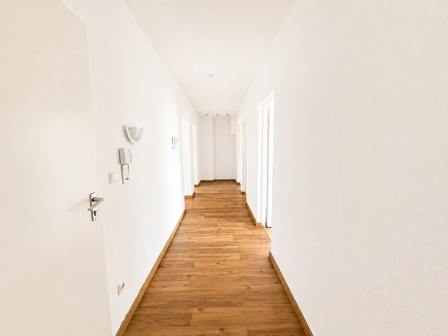 Bright, empty hallway with white walls and a light brown wood floor. A door is on the left, and doorways are visible at the end.