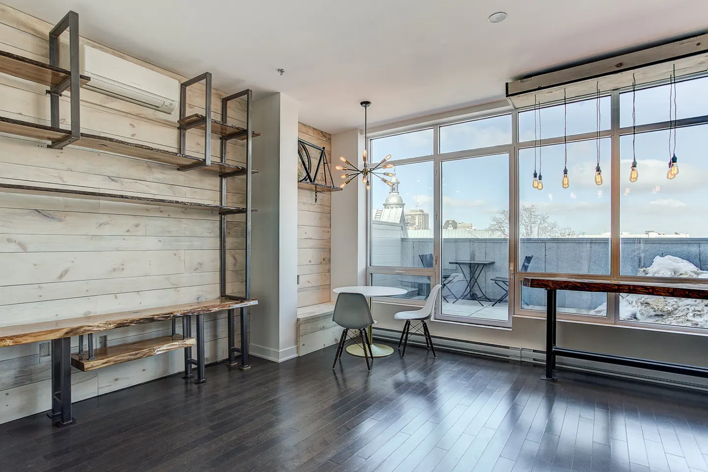 Bright, modern apartment interior with wood floors, white walls, and large windows overlooking a city view. Shelving and tables are made of wood and metal.