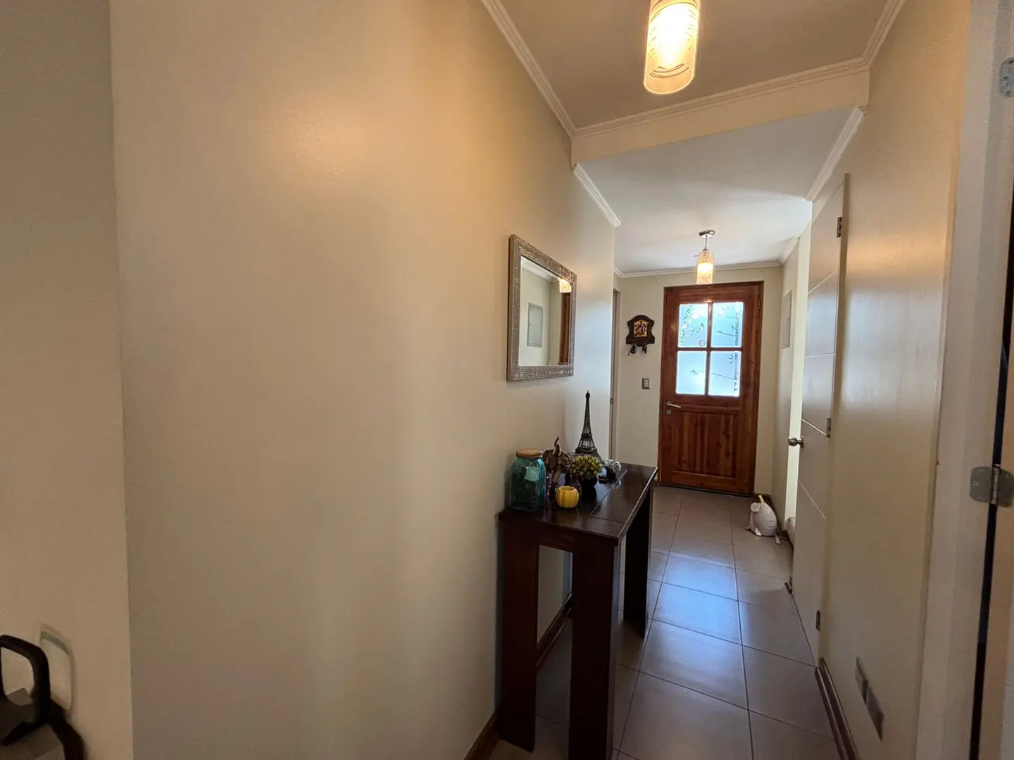 Hallway with beige walls, a dark wood table with decor, and a mirror. A wood door is at the end of the hall.