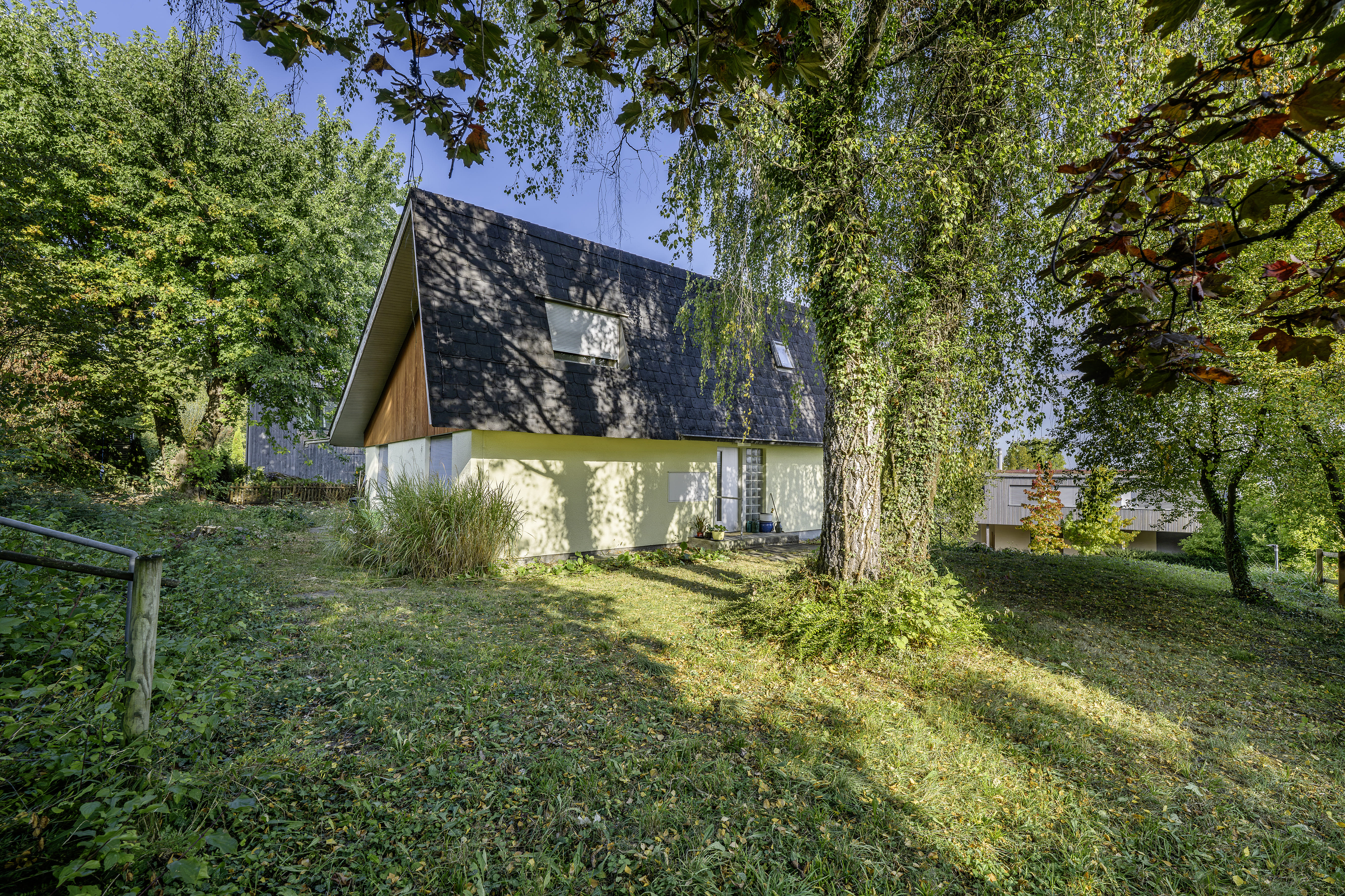 A yellow A-frame house with a dark shingled roof sits in a grassy, tree-filled yard on a sunny day. [AI-generated text]