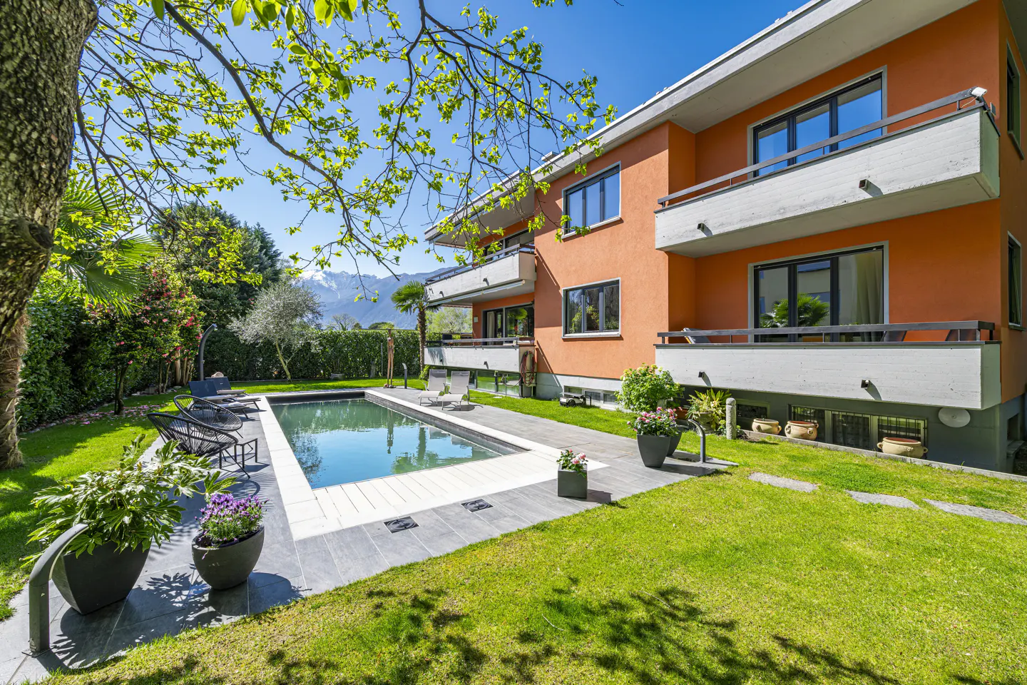 Orange building with balconies overlooks a pool and green lawn. Trees and mountains are in the background.