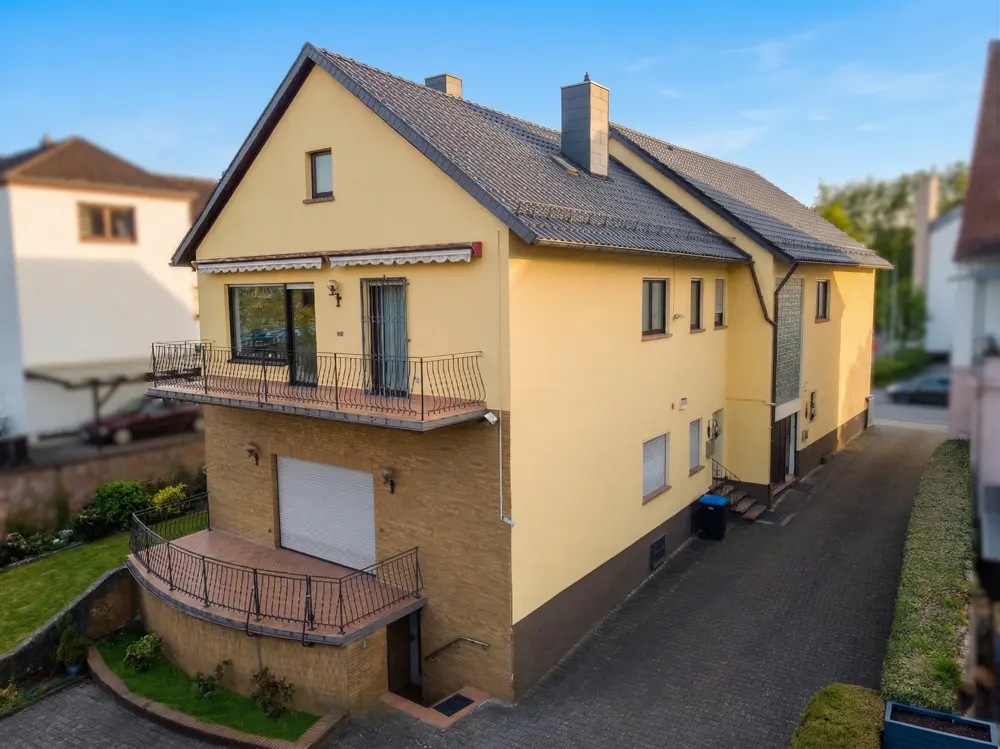 Two-story yellow house with gray roof, balconies, and a driveway. The lower level is brick.