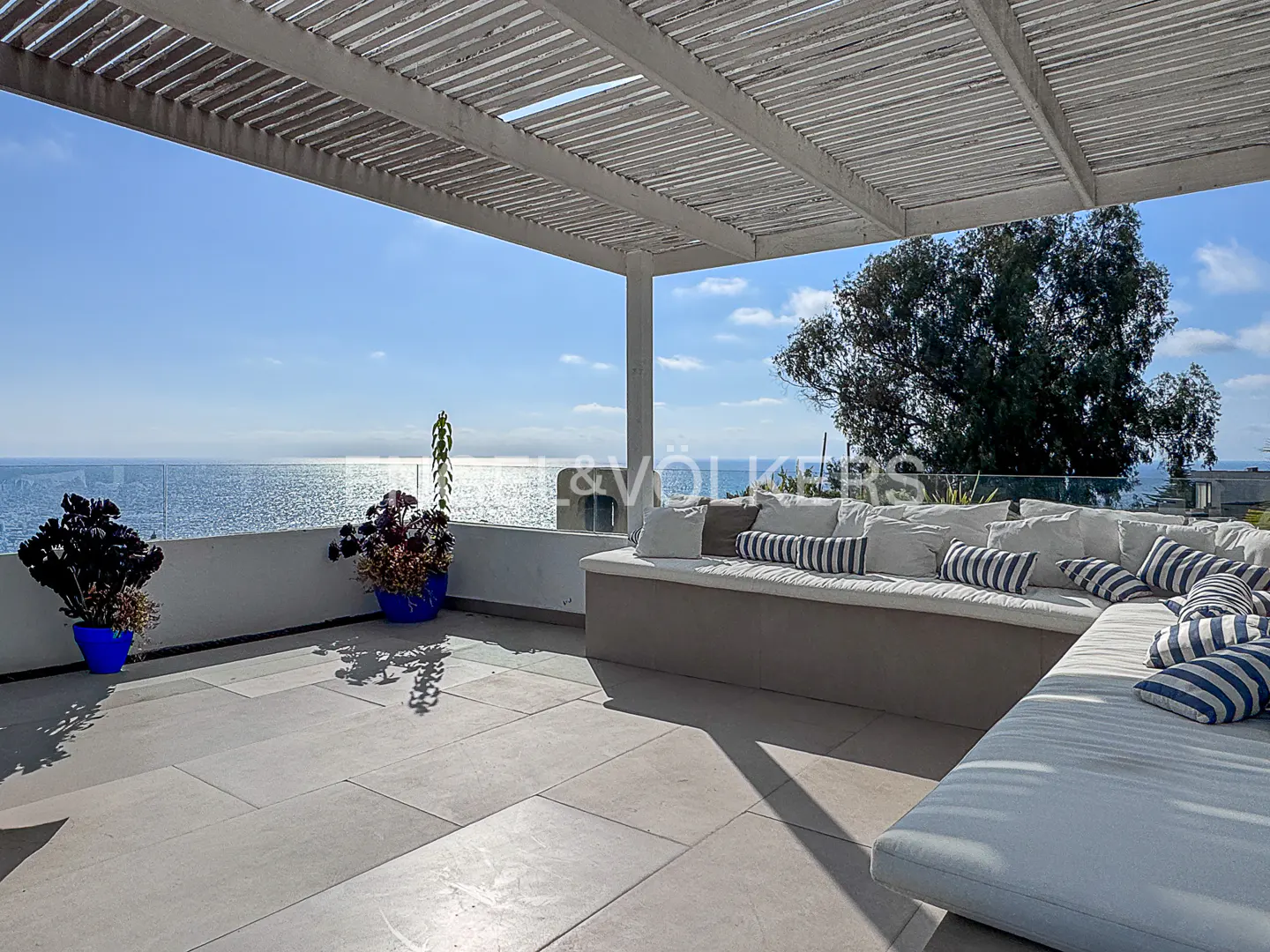 Outdoor patio with ocean view, featuring a white pergola, stone flooring, and a built-in sofa with white and blue striped pillows.