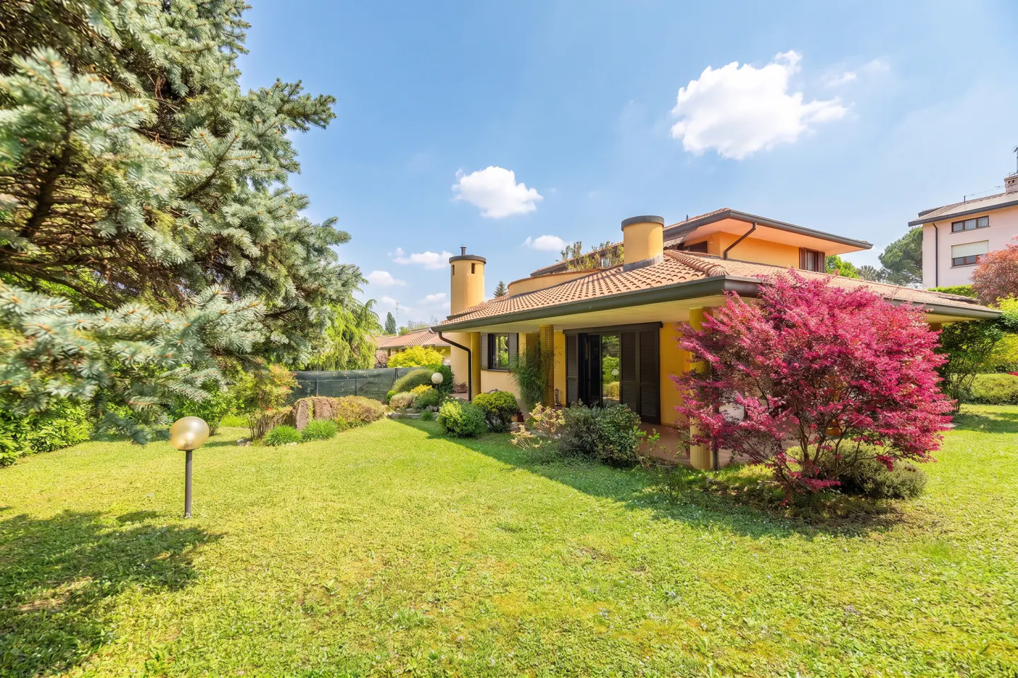 A yellow house with a red-tiled roof sits on a green lawn with trees and bushes under a blue sky.
