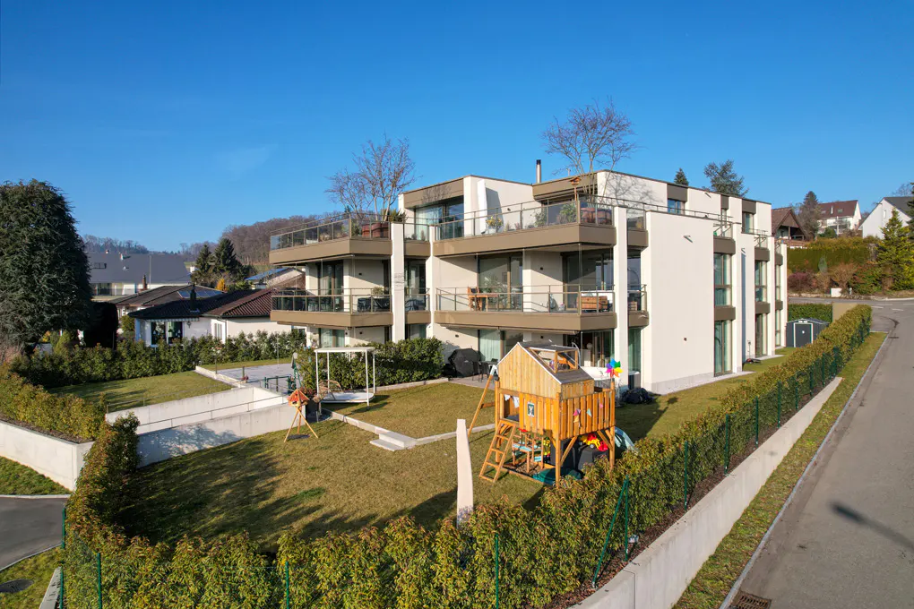 Modern white apartment building with brown balconies and a green lawn with a wooden playset.