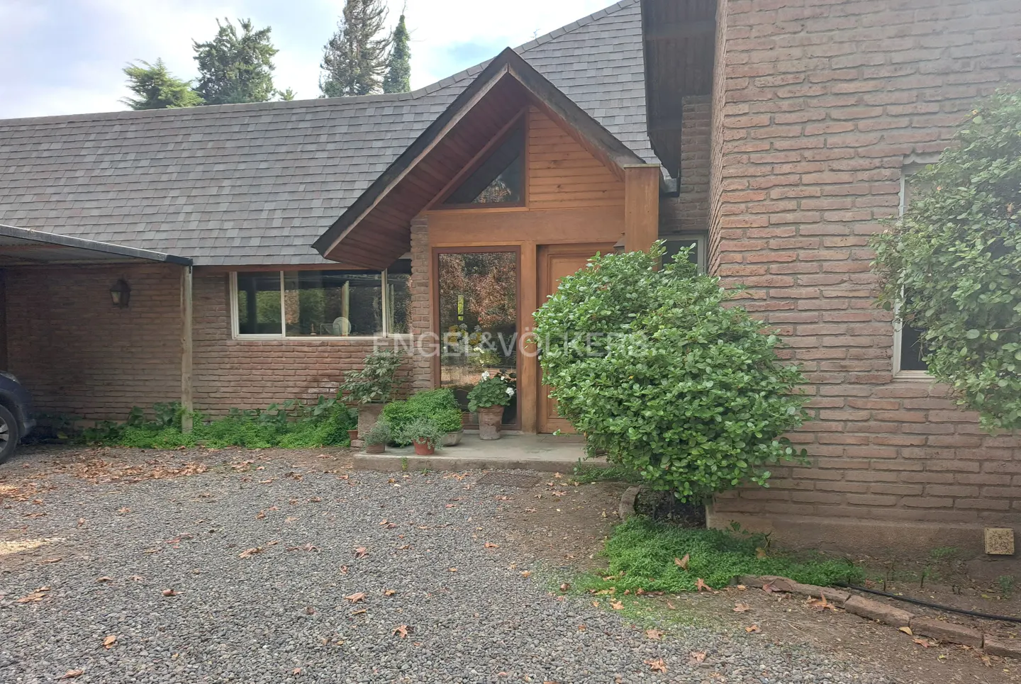 Exterior view of a brick house with a gray shingled roof and a gravel driveway. The entrance has wooden accents and potted plants.