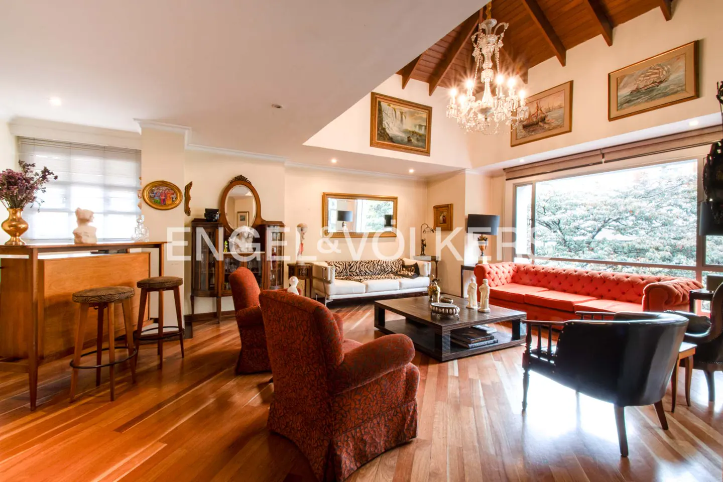Spacious living room with hardwood floors, a crystal chandelier, a red tufted sofa, and patterned armchairs.