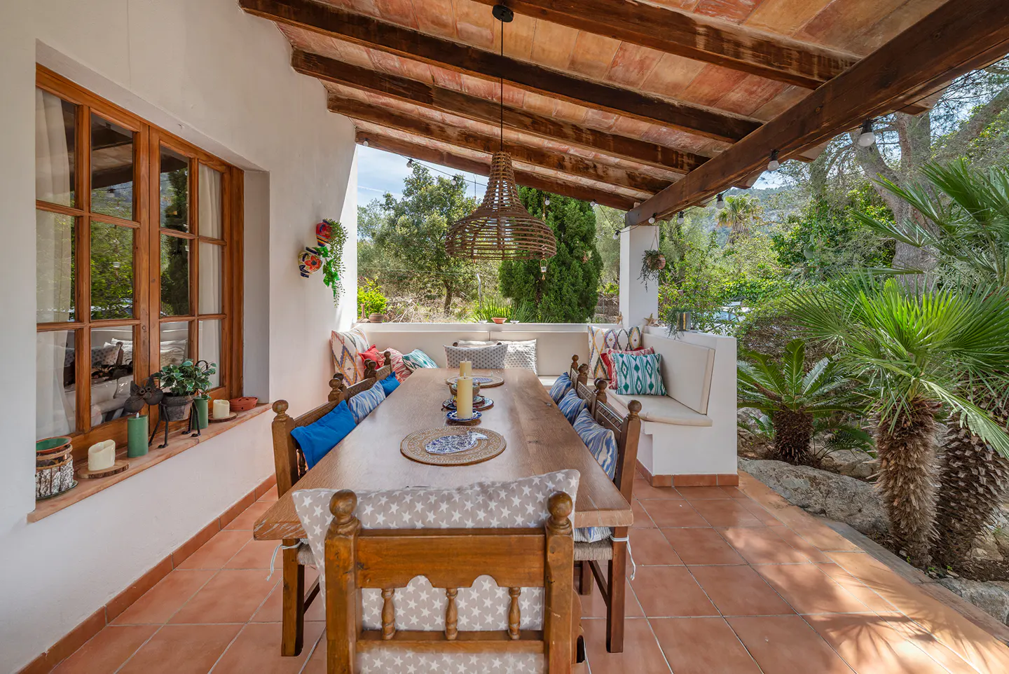 Outdoor dining area with a long wooden table, chairs, and cushions under a covered patio with a view of lush greenery.