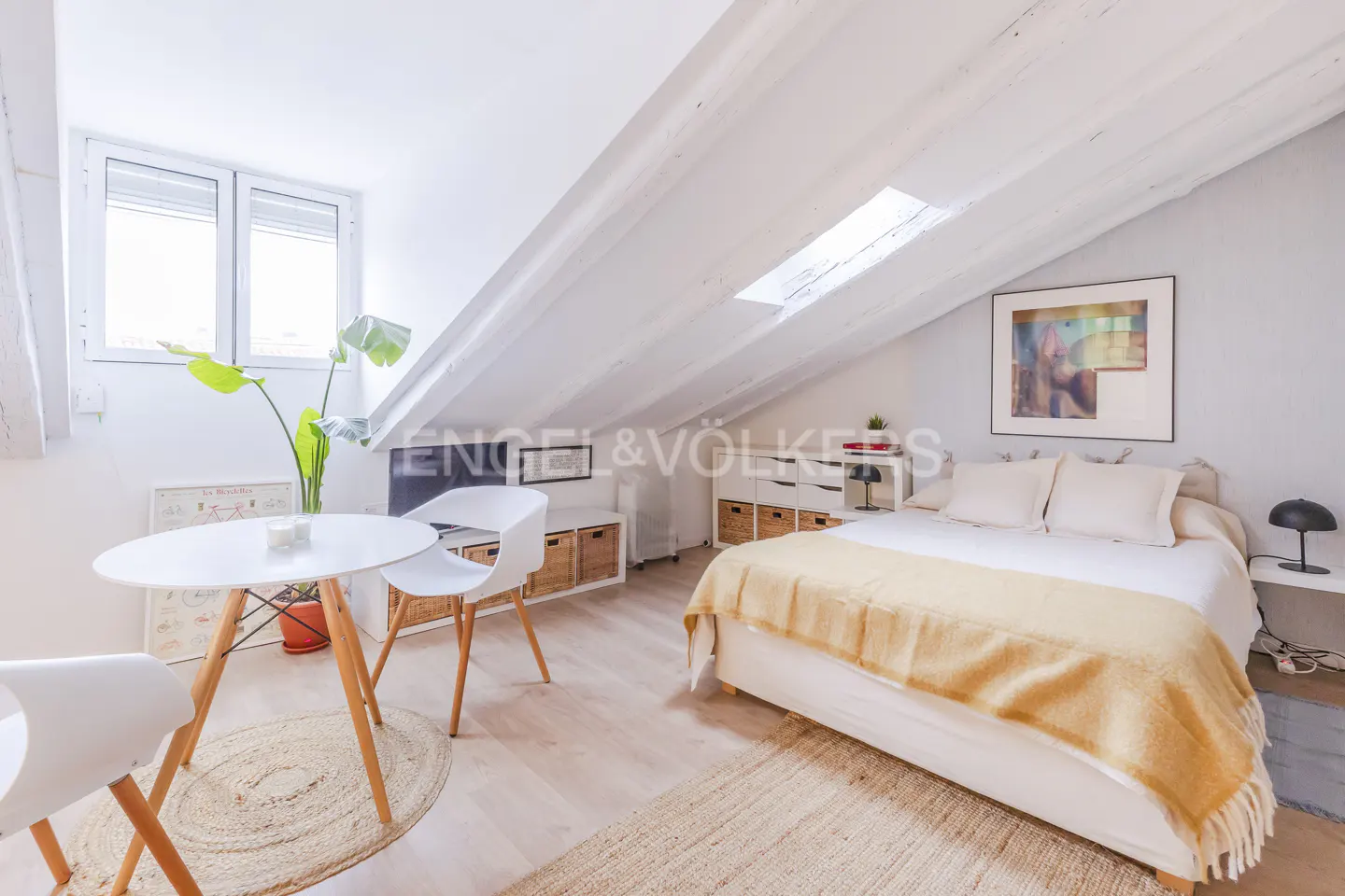 Attic bedroom with white walls, exposed beams, and a skylight. A bed, table, chairs, and TV are arranged in the space.