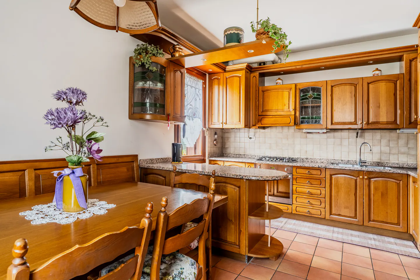 A warm kitchen with wood cabinets, granite countertops, and terracotta tile flooring. A dining table with purple flowers sits in the foreground.