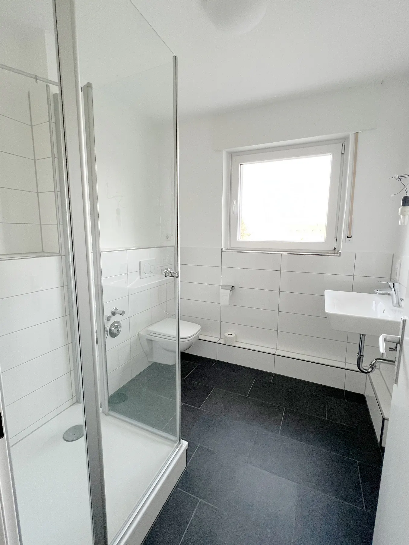 Bright bathroom with black tile floor, white walls, toilet, sink, and glass shower stall. A window provides natural light.