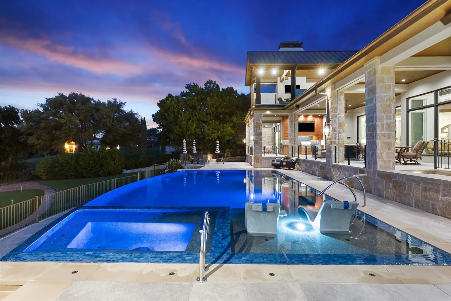 Exterior view of a luxury home with a blue-lit pool and spa at dusk. Stone columns support a covered patio with seating. Trees and a colorful sky are in the background.