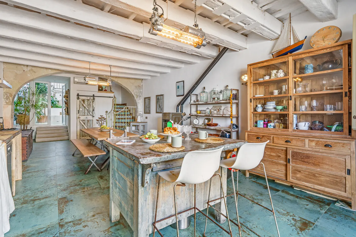 Rustic kitchen with white beamed ceiling, blue floor, and wooden cabinets. An island with white stools sits in the center.