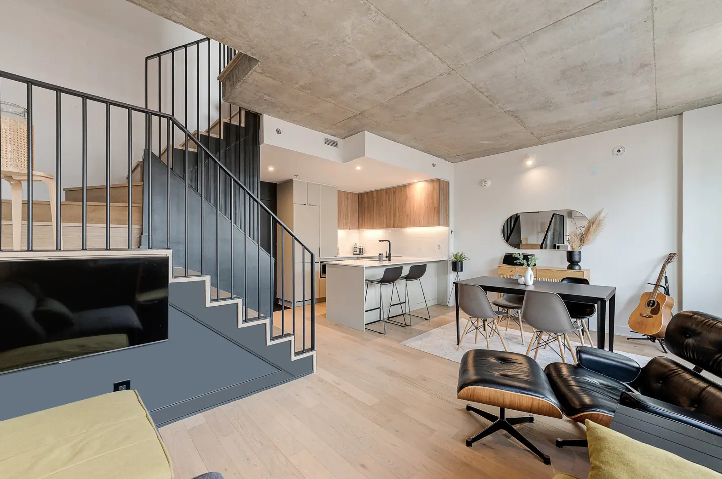 A modern apartment interior with a staircase, kitchen island, dining table, and black leather lounge chair.