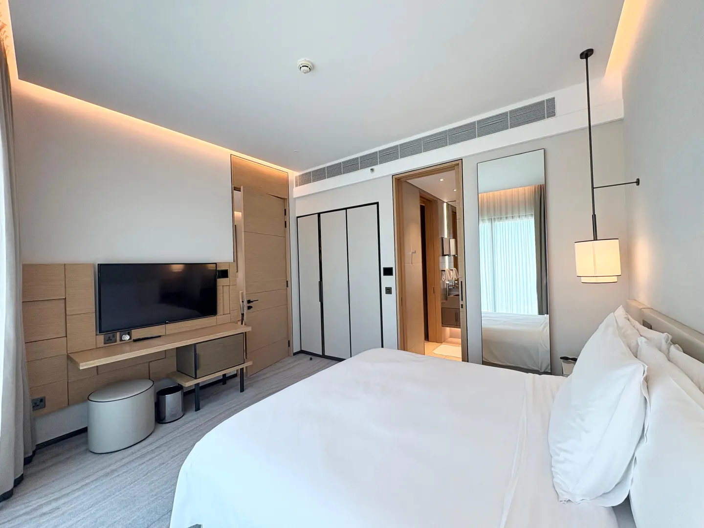 Modern hotel bedroom with a white bed, wall-mounted TV, light wood accents, a tall mirror, and warm recessed lighting.