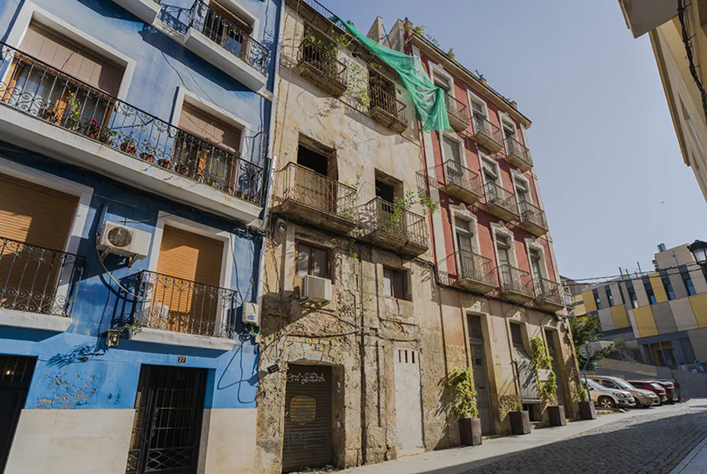 Street view of colorful, old buildings with balconies. Blue, beige, and red facades. Cobblestone street and parked cars in the background.