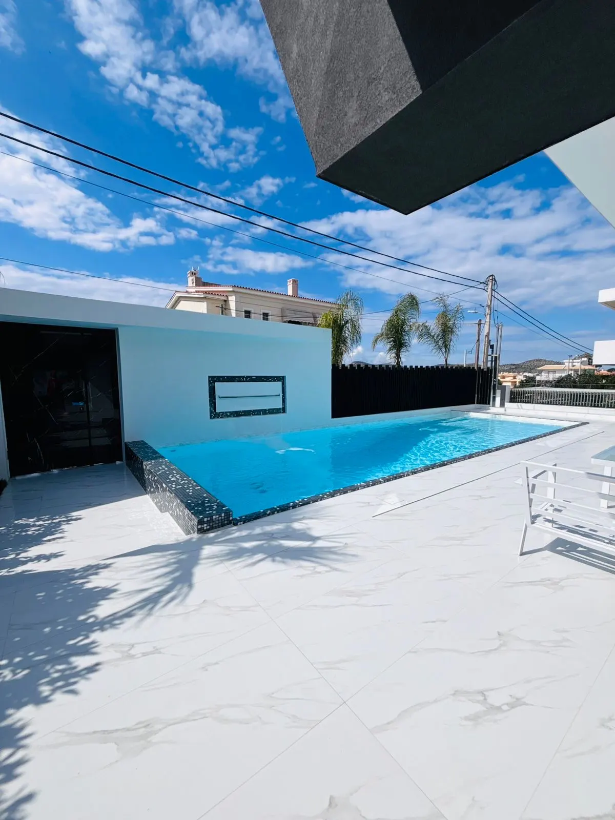 A modern home's outdoor pool area with white tile, blue water, and a black accent wall under a partly cloudy sky.