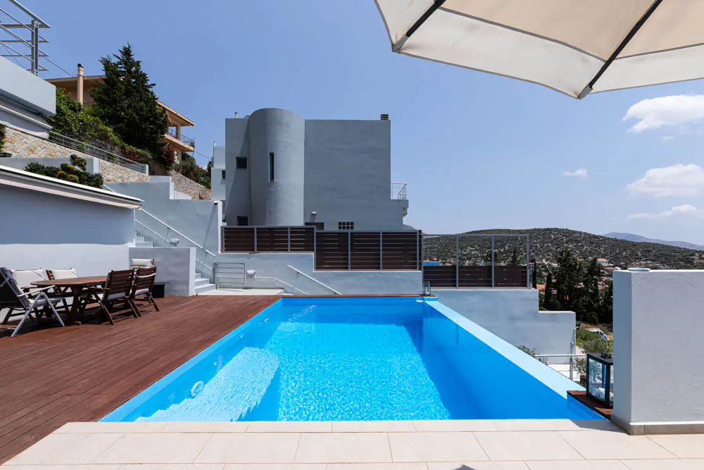 A bright blue pool is centered in front of a modern white house with a brown deck and table with chairs.