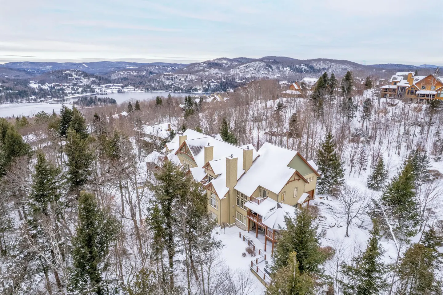 Aerial view of a tan house with snow-covered roof, surrounded by trees and a snowy landscape. Mountains and a lake are in the background.