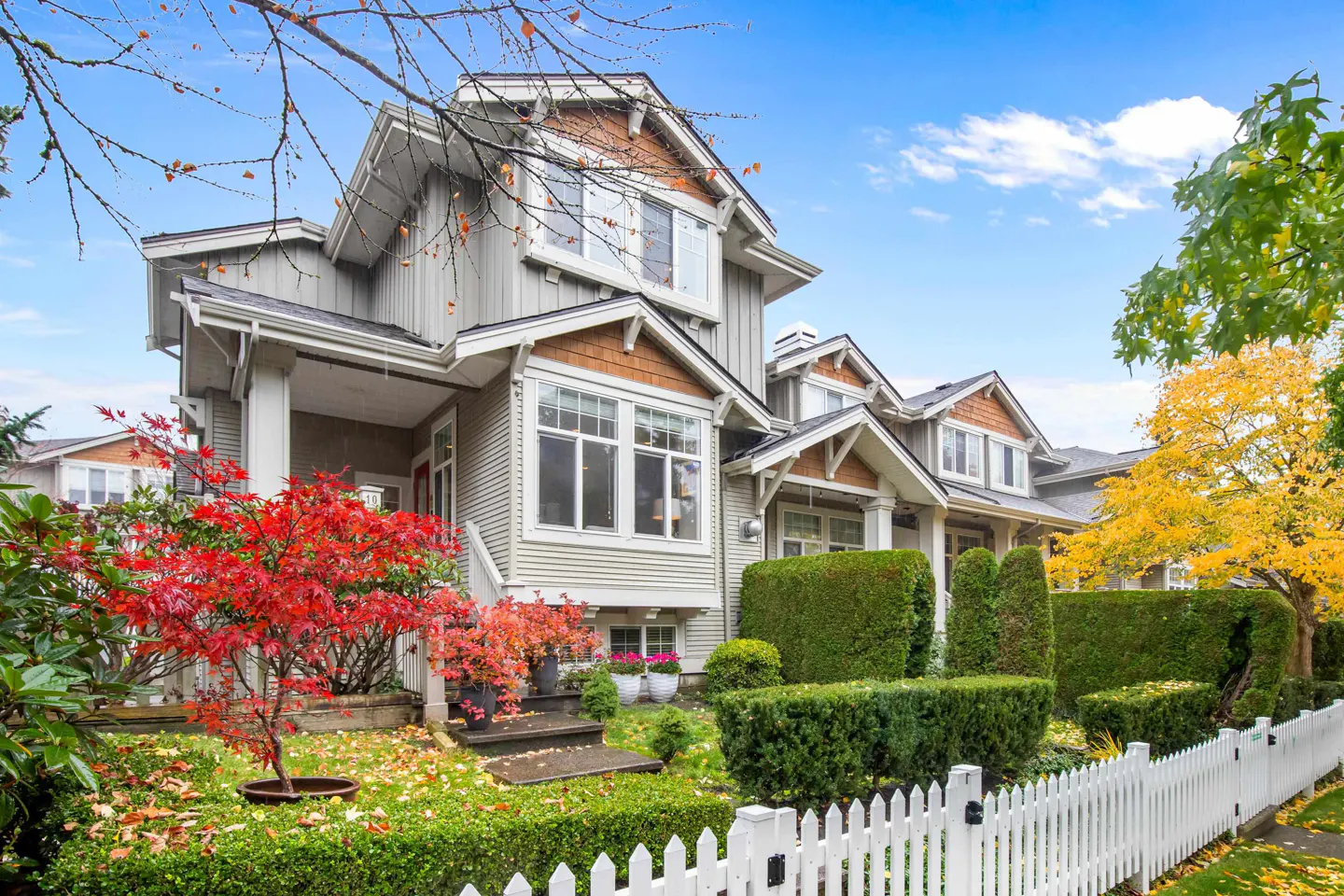 A two-story gray house with white trim and a red maple tree in the front yard. A white picket fence lines the property.