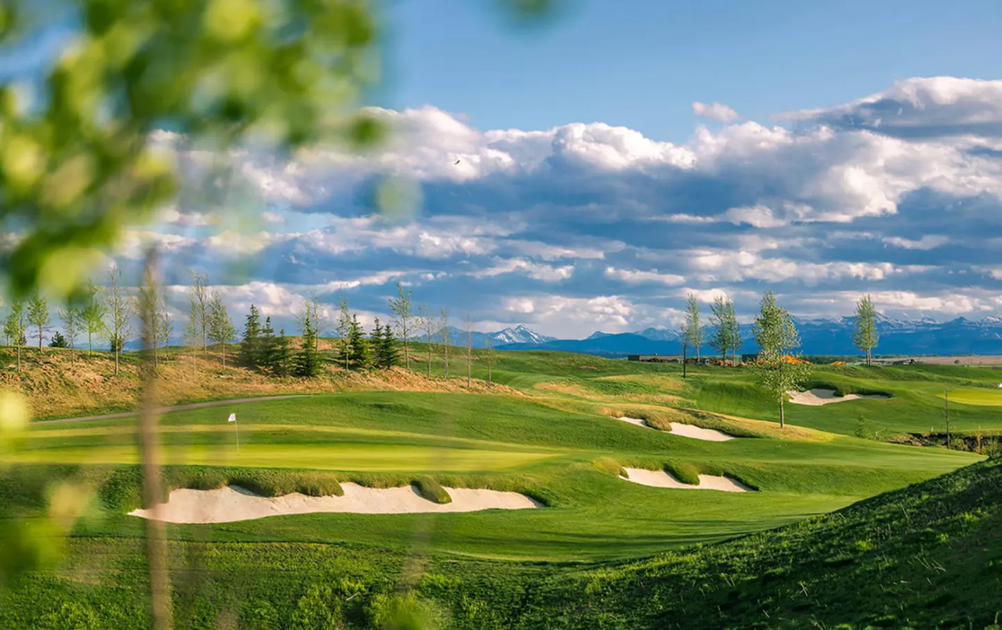 Scenic view of a green golf course with sand traps under a cloudy blue sky and distant snow-capped mountains.