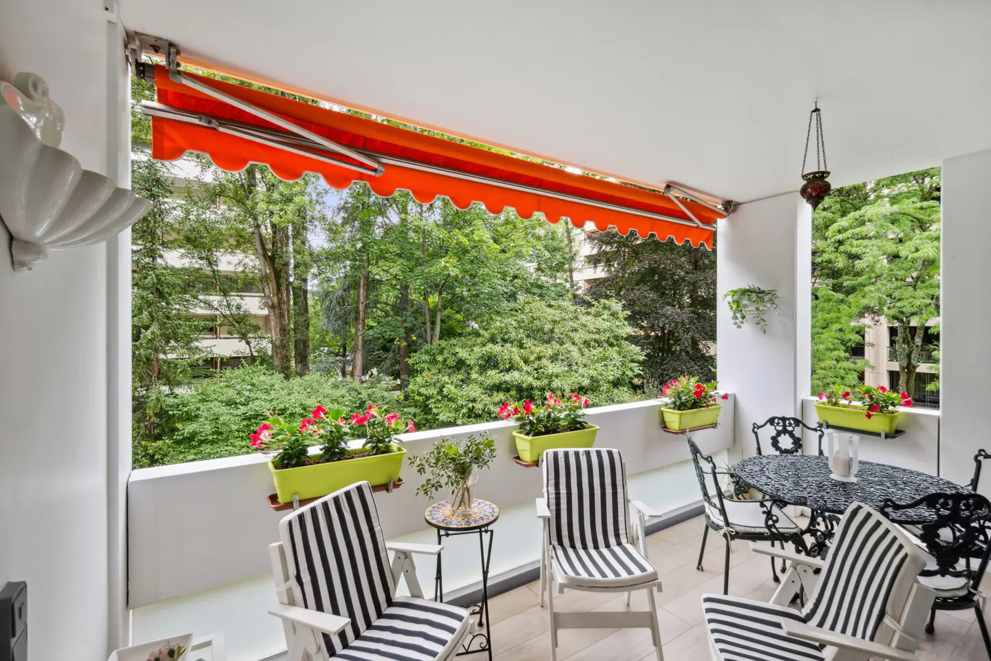 A bright balcony with black and white striped chairs, a black table, and green flower boxes overlooking lush greenery. An orange awning provides shade.