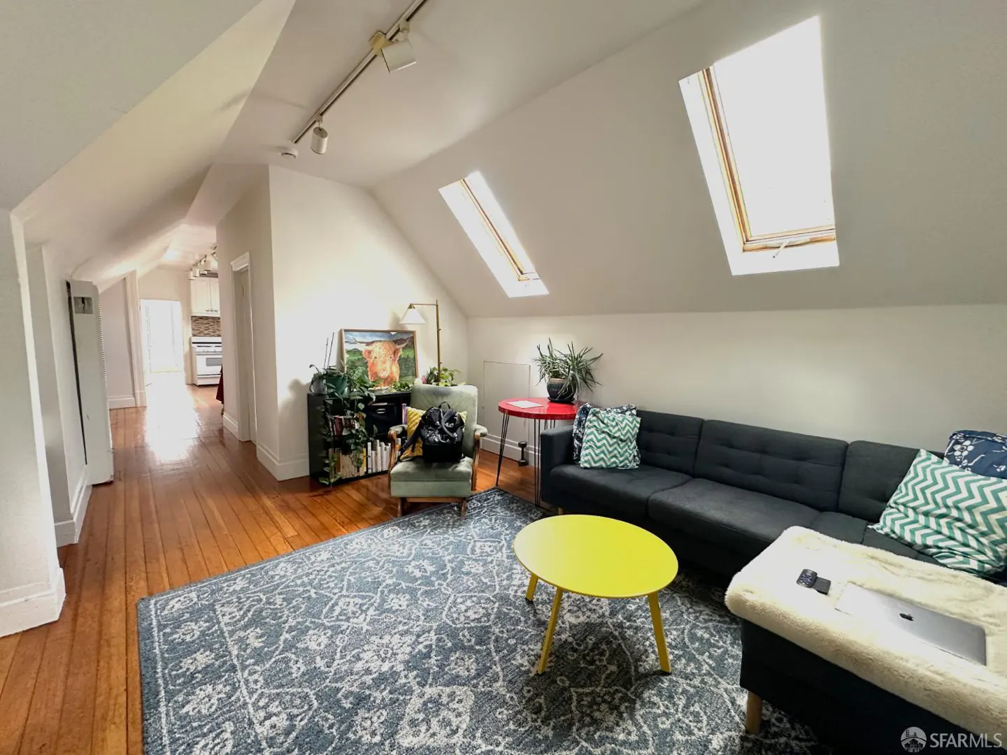 Attic living room with skylights, hardwood floors, and a blue patterned rug. A gray sofa, yellow table, and green chair furnish the space.