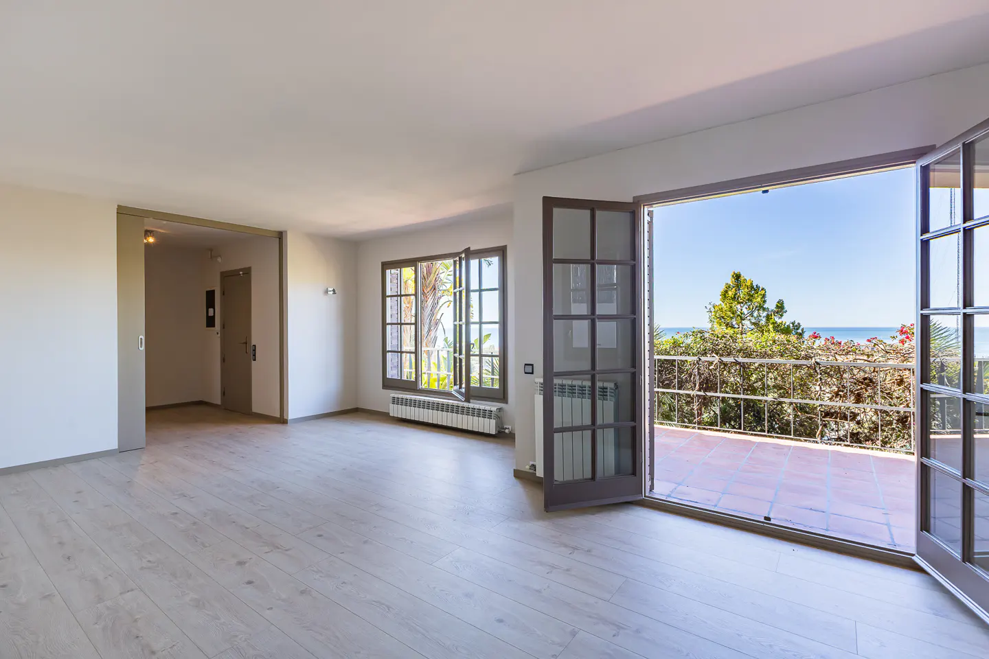 Bright, empty room with light wood floors, white walls, and open doors to a balcony with ocean views.