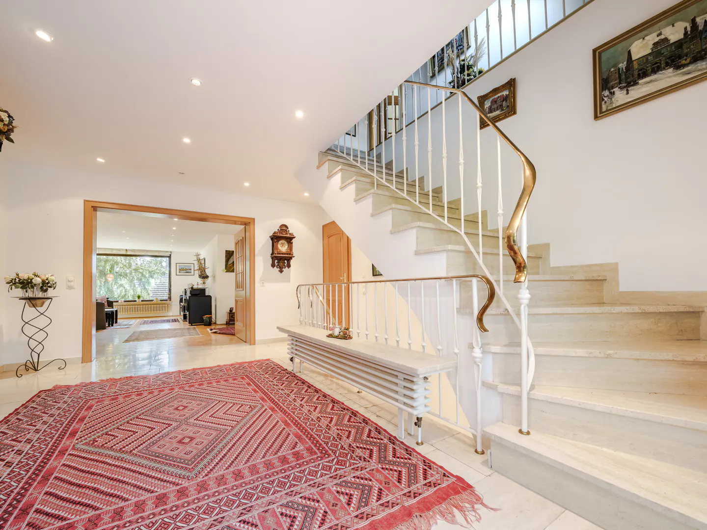 Bright foyer with marble stairs, white railing, and gold accents. A red patterned rug covers the floor. A doorway leads to a living room.